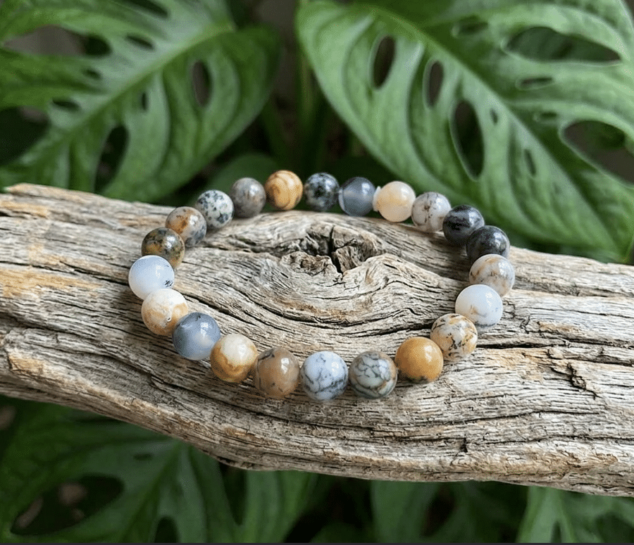 Un bracelet agate dendrite fait de perles de pierres rondes de 8 millimètres est posé sur un morceau de bois flotté gris et texturé. Les pierres ont différentes couleurs, du blanc cassé au gris en passant par le jaune moutarde et le bleu poudré, avec des motifs et des inclusions uniques. En arrière-plan, on voit une plante monstera verte luxuriante avec ses feuilles palmées distinctives.