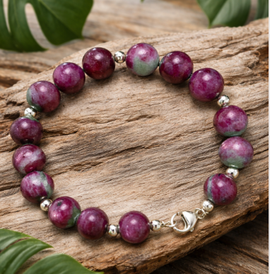 Bracelet en perles rondes violettes de rubis zoïsite naturelle, monté avec de petites perles en argent 925 et un fermoir argent 925, posé en cercle. Il repose sur une planche de bois flotté au relief marqué, donnant un aspect naturel et rustique. Autour, de grandes feuilles vertes de monstera encadrent la scène et apportent une ambiance tropicale douce.