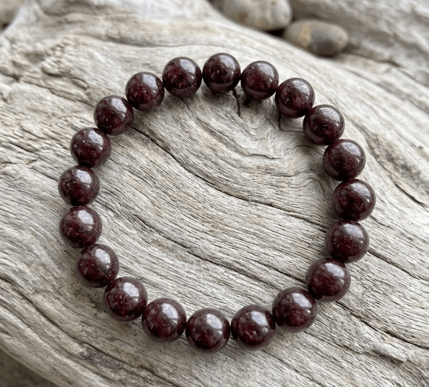Bracelet en perles de grenat de 8 mm sur bois flotté Ce bracelet est composé de 22 perles rondes plus volumineuses (diamètre 8 mm), aux nuances rouge grenat et bordeaux très sombres et profondes. Il repose à plat sur un morceau de bois flotté gris clair, aux fibres très marquées, qui met en valeur la brillance et le galbe des pierres.