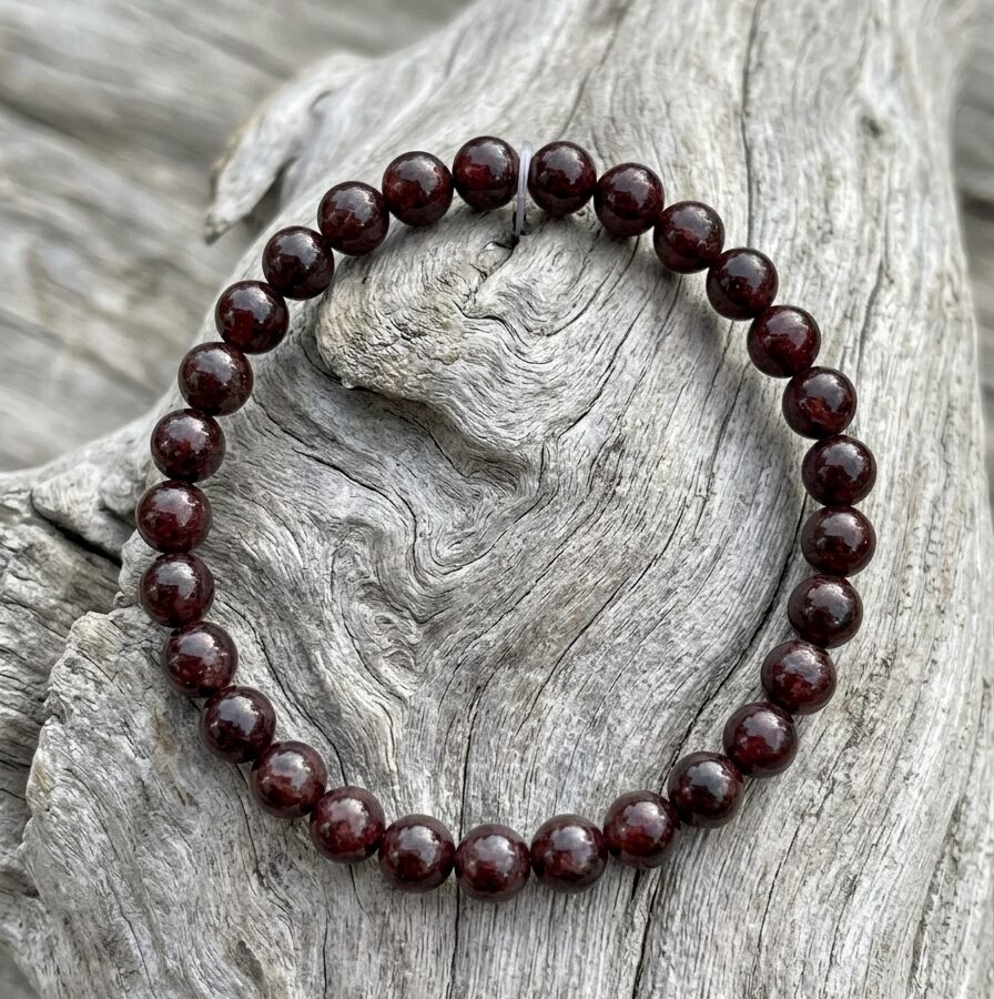 Bracelet en pierres rondes sur bois de grève Ce bracelet présente une enfilade de perles de pierres rondes et lisses, d'une couleur rouge bordeaux profond et intense. Il est posé sur un morceau de bois de grève, dont la surface est usée et patinée, de couleur gris clair, avec un veinage marqué.