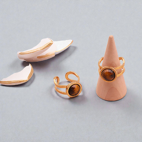 Deux bagues en œil de tigre style Azélis doré sur un fond clair et lumineux : une bague posée à plat et l’autre sur un petit présentoir en forme de cône, pierre centrale de 10 mm, monture en acier inoxydable professionnel, bagues artisanales, reflets dorés et bruns, bijou élégant et protecteur.