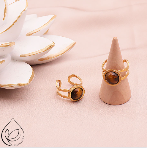 Deux bagues en œil de tigre style Azélis doré sur un fond clair et lumineux : une bague posée à plat et l’autre sur un petit présentoir en forme de cône, pierre centrale de 10 mm, monture en acier inoxydable professionnel, bagues artisanales, reflets dorés et bruns, bijou élégant et protecteur.