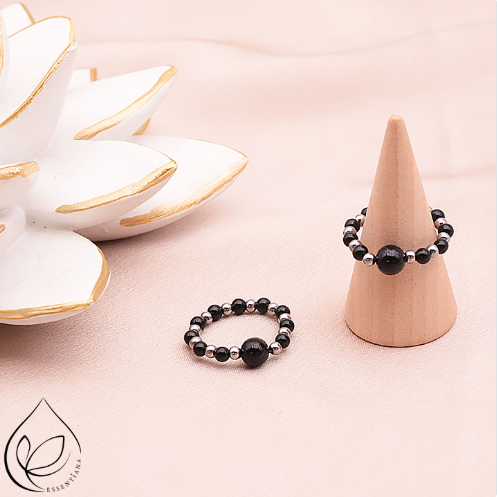 Deux bagues en onyx et hématite sur un fond clair et lumineux : une bague posée à plat et l’autre sur un petit présentoir en forme de cône, perles de 3 à 6 mm, bagues artisanales, pierres naturelles qualité A, couleurs noir profond et argenté, bijou moderne et raffiné.