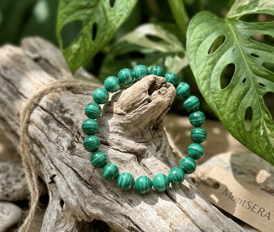 Un bracelet malachite de perles rondes vert intense, avec des marbrures concentriques noires, est délicatement posé sur un morceau de bois flotté texturé et blanchi. L'arrière-plan est composé de feuilles vertes et blanches de Monstera, créant un décor naturel et végétal pour le bijou.