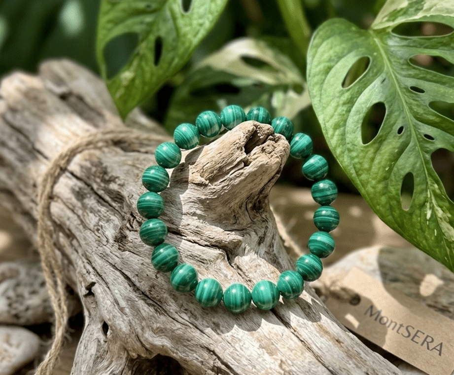 Un bracelet malachite de perles rondes vert intense, avec des marbrures concentriques noires, est délicatement posé sur un morceau de bois flotté texturé et blanchi. L'arrière-plan est composé de feuilles vertes et blanches de Monstera, créant un décor naturel et végétal pour le bijou.