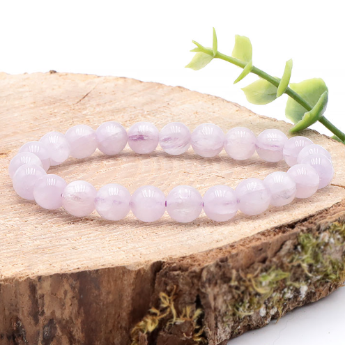 Un bracelet circulaire en Kunzite aux perles rondes et lisses, affichant des nuances douces de rose pâle et de lilas, est posé à plat au centre de grandes feuilles de Monstera vertes et brillantes. Le contraste entre les tons pastels des pierres et le vert profond du feuillage exotique crée un visuel naturel et apaisant.