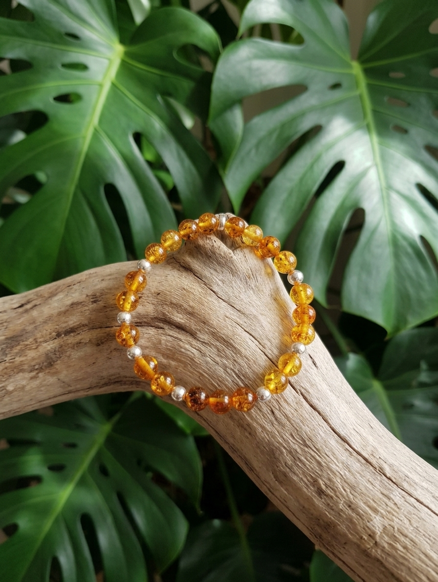 Un bracelet de perles d'ambre rondes et de perles d'argent alterne sur un fil transparent. Le bracelet repose sur une grosse branche de bois flotté rugueuse et texturée. L'arrière-plan est flouté et montre de grandes feuilles vertes de Monstera.