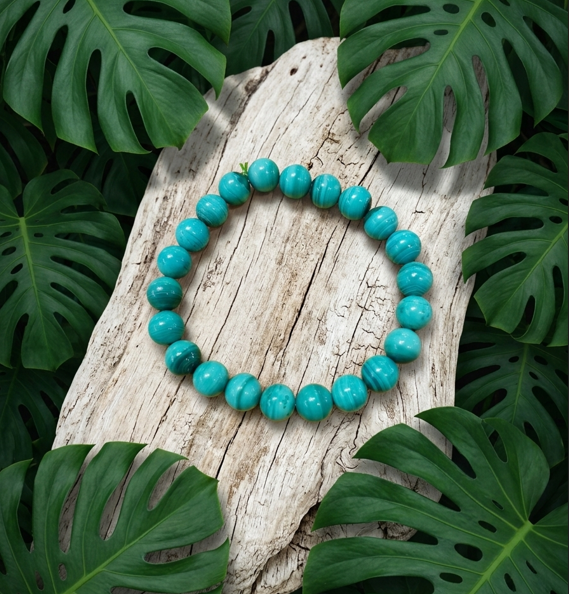 bracelet en perles rondes de malachite verte aux marbrures turquoise, disposé à plat au centre d'un large morceau de bois flotté blanchi et texturé. L'ensemble est entouré en premier plan et en arrière-plan par de grandes feuilles vertes et découpées de Monstera, créant une ambiance végétale et naturelle.
