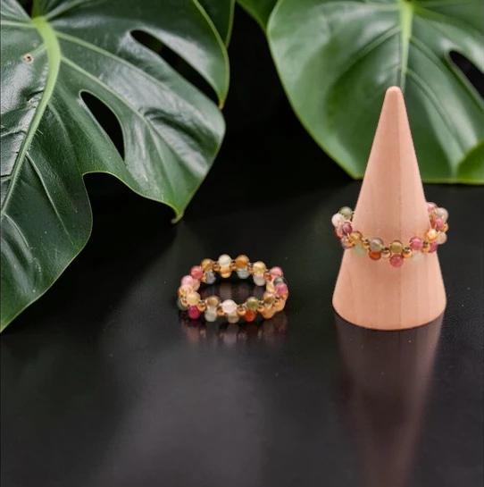 Deux bagues en tourmaline multicolore présentées sur un fond clair et lumineux : une bague posée à plat et l’autre sur un petit présentoir en forme de cône, pierres naturelles de qualité AA+, perles de 3 mm, bijou artisanal fait main, éclat et couleurs bleu, doré, jaune, rose et vert.