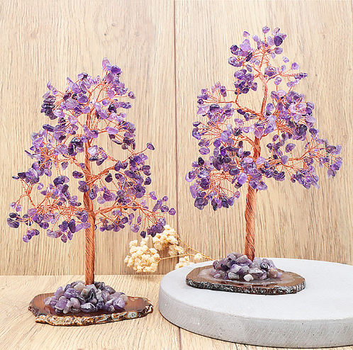 Cette image présente deux arbres de vie améthyste artisanaux, d'environ 20 cm de hauteur, avec des troncs et des branches en fil de cuivre torsadé. Leur feuillage est constitué d'une multitude de petits fragments d'améthyste violette, offrant des reflets éclatants et variés. Chaque arbre repose sur une base naturelle en agate d'une couleur plus sombre. À l'arrière-plan, un mur en bois clair crée une atmosphère naturelle et chaleureuse.