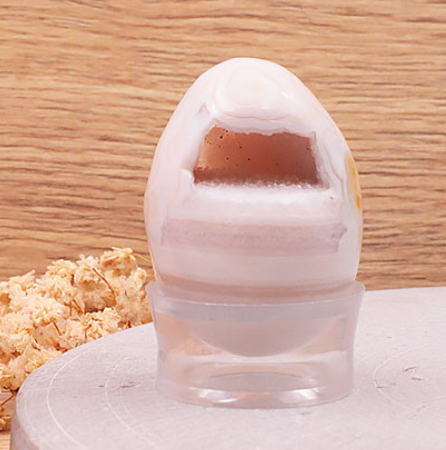 Cette image présente un œuf décoratif en agate rose clair, mesurant environ 40 mm de hauteur. L'œuf est doté d'une ouverture taillée qui révèle une géode interne étincelante et cristallisée. Il est présenté sur un petit socle en acrylique transparent, mais veuillez noter que ce socle n'est pas inclus lors de l'achat. L'arrière-plan présente un fond boisé chaleureux et un bouquet de fleurs séchées.