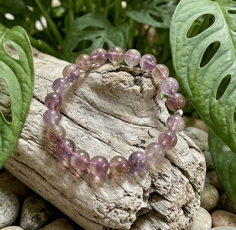 Un bracelet en perles de pierre de ton violet transparent et nuances ambrées repose sur un morceau de bois flotté strié. Le bois est posé sur un lit de galets gris et bruns. En arrière-plan, de grandes feuilles vertes perforées de Monstera encadrent la scène. L'ambiance générale est naturelle et végétale.