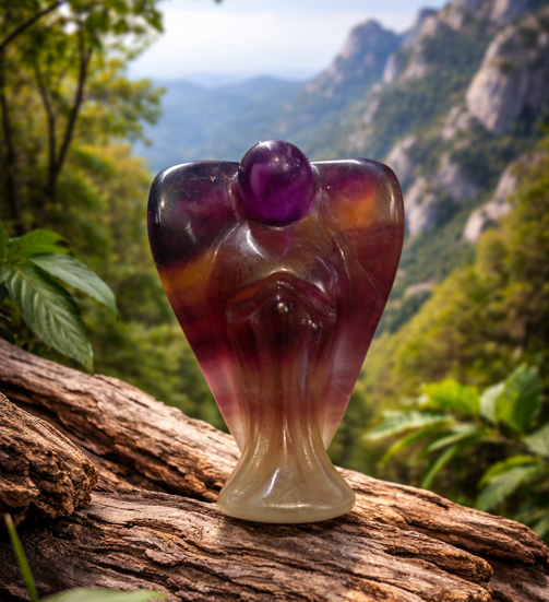 Un petit ange sculpté dans une pierre polie est placé au centre de l’image. Il tient debout, avec des formes douces et arrondies. La pierre présente de belles nuances de couleurs : du violet foncé au niveau de la tête, qui est en forme de petite boule, puis des tons violets, orangés et translucides qui descendent vers le corps, jusqu’à une base plus claire et presque blanche. L’ange repose sur du bois flotté, aux textures rugueuses et craquelées, de couleur brun clair. Autour, on distingue des feuilles vertes floues, qui donnent une impression de nature. En arrière-plan, le paysage est également flou : on aperçoit des montagnes rocheuses et verdoyantes sous un ciel lumineux, ce qui crée une ambiance naturelle et apaisante. La lumière est douce et met en valeur la transparence et les reflets de la pierre, donnant à l’ensemble une sensation chaleureuse et harmonieuse.