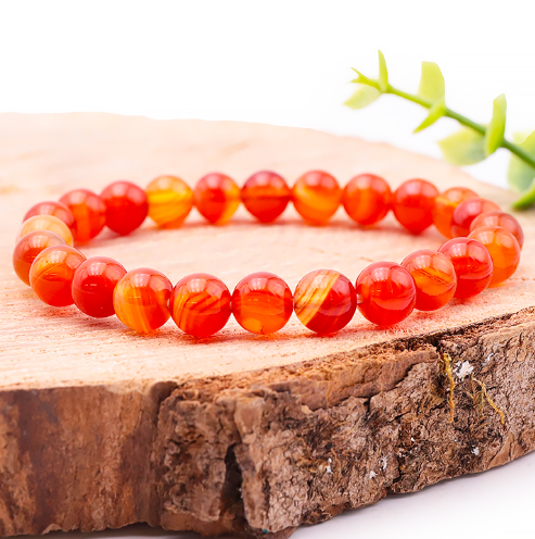 Ce bracelet circulaire est composé de perles d'agate aux tons orange vif et rouge brique, présentant des bandes zébrées naturelles plus claires. Les pierres polies et brillantes reposent sur un support en bois rustique, créant un contraste chaud et dynamique. Une petite touche végétale verte en arrière-plan souligne l'aspect naturel et authentique de l'ensemble.