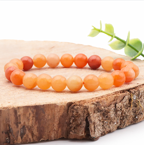 Ce bracelet délicat en Aventurine orange se compose de petites perles rondes aux nuances vitaminées allant du jaune orangé au corail doux. Il est photographié de profil sur un socle en bois brut dont l'écorce est apparente, créant un contraste entre la pierre lisse et la texture rugueuse. L'image, lumineuse et épurée, est rehaussée par une petite branche verte en arrière-plan.