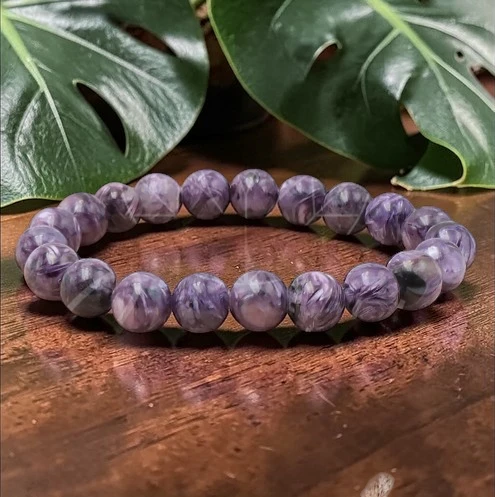 Le bracelet est un bijou en pierres naturelles de charoïte, présenté sous forme d'une boucle continue de perles parfaitement sphériques et lisses. Aspect des pierres : Chaque perle est unique, offrant une robe violette intense qui varie du parme clair au prune profond. On y distingue des motifs tourbillonnants et des veines nacrées (blanches et grises) qui donnent un aspect soyeux et marbré à la pierre. Quelques inclusions plus sombres, presque noires, ajoutent du caractère à l'ensemble. Finition et texture : Les perles sont polies pour obtenir un éclat brillant et un toucher très doux. Elles captent la lumière, ce qui accentue la profondeur de leurs nuances violettes. Montage : Le bracelet est composé d'une vingtaine de perles de taille régulière (environ 8 mm), montées sur un fil élastique invisible qui permet de l'ajuster facilement au poignet. L'ambiance de l'image : Le bracelet occupe le centre de la photo, posé sur un bois sombre et chaleureux, avec des feuilles de Monstera en arrière-plan qui soulignent son origine naturelle et minérale.