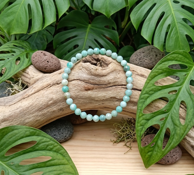 Un bracelet de perles rondes en pierre bleu-vert clair est suspendu au centre, devant une branche de bois flotté sculptée. Il est entouré de roches volcaniques grises et rouges et de mousse. De grandes feuilles vertes de Monstera aux motifs de découpes et de trous encadrent la scène. L'ensemble repose sur un plateau en bois clair.