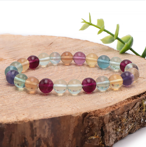 Ce bracelet en fluorine multicolore se compose de perles translucides de 8 mm aux nuances éclatantes de violet, bleu, jaune et vert. Les pierres, à l'aspect vitreux et lumineux, sont présentées sur un support en bois naturel qui souligne leur transparence et leur éclat.