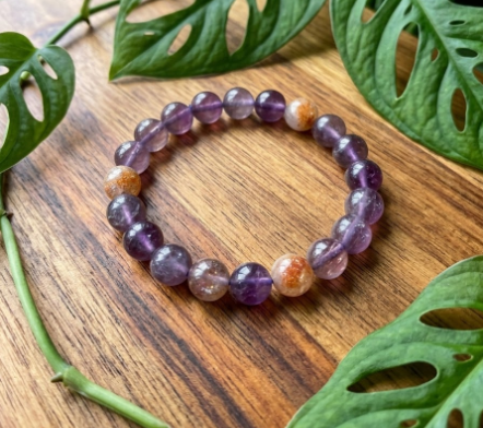 Ce bracelet en fluorine violette et pierre de soleil se compose de perles translucides aux teintes améthyste, illuminées par des perles orangées scintillantes. Il repose sur un bois de chêne chaleureux, entouré de feuilles de Monstera d'un vert vif qui soulignent son éclat naturel. L'ensemble dégage une atmosphère à la fois apaisante et solaire.