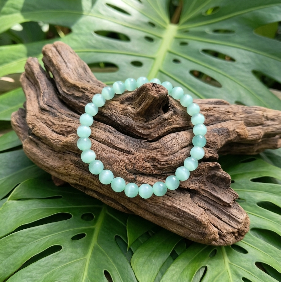 Un bracelet de perles rondes vert pâle et brillantes est posé sur un morceau de bois flotté texturé. Le tout repose sur de grandes feuilles de monstera d'un vert vif, créant un décor naturel et harmonieux.