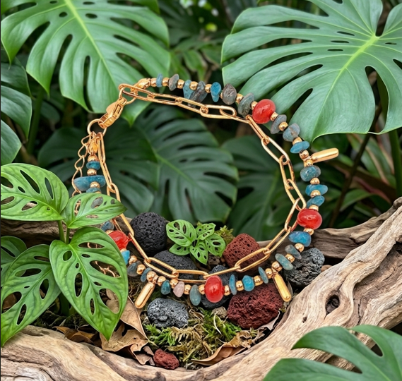 Un bracelet à double rangée est présenté dans un décor naturel tropical. Il se compose d'une fine chaîne dorée à maillons allongés et d'un second rang orné de petites pierres bleu turquoise et de perles rouges lisses, agrémenté de petits pendentifs cylindriques dorés. Le bijou repose sur un morceau de bois flotté creusé, entouré de mousse, de petites roches volcaniques noires et rouges, le tout encadré par de larges feuilles vertes de Monstera.