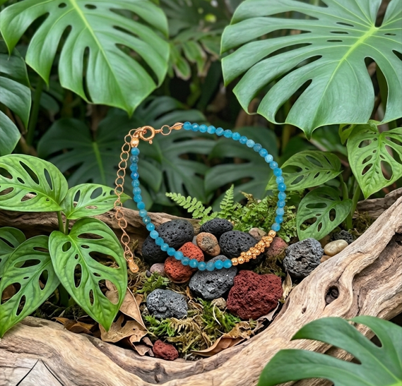 Un bracelet fin repose dans une coupelle en bois naturel sculpté. Il est composé de petites perles bleu vif et d'un fermoir cuivré. Le bijou est entouré de roches volcaniques noires et rouges et de mousse. De grandes feuilles vertes de Monstera encadrent la scène en arrière-plan.