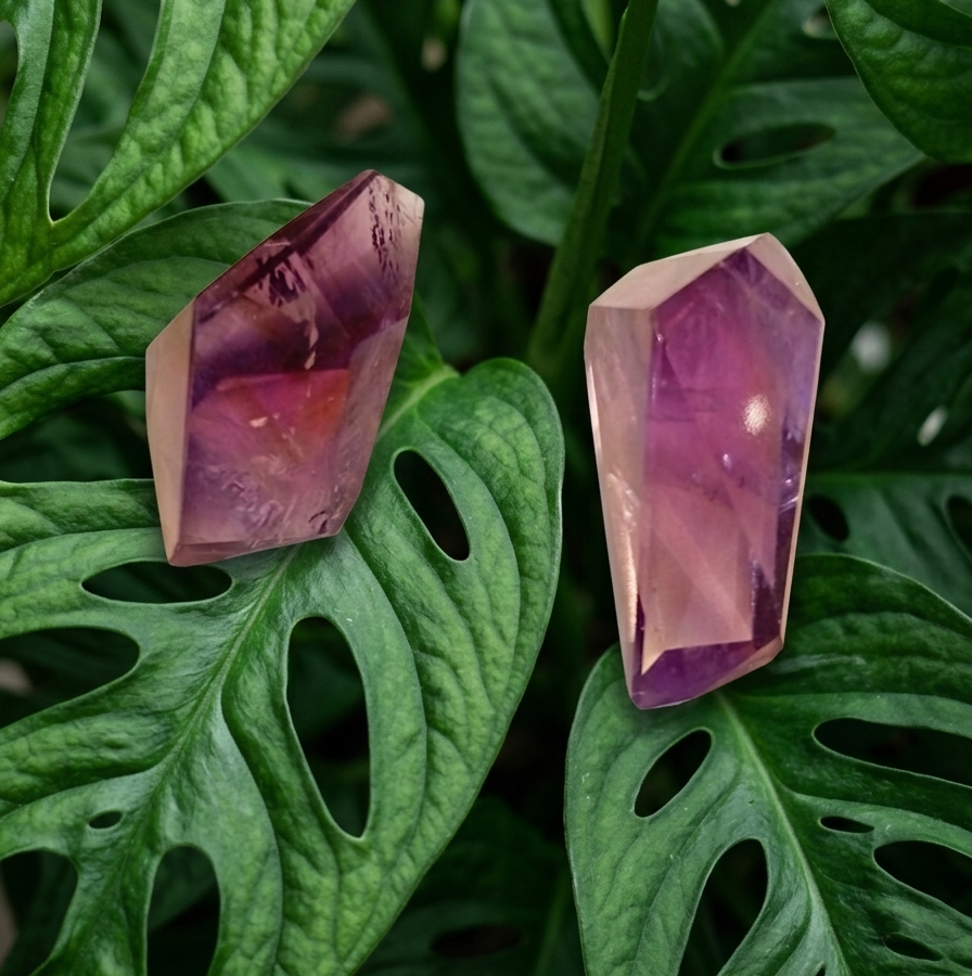Deux pierres d'améthyste polies et taillées en forme de pointes reposent sur de grandes feuilles de Monstera vert vif. Le feuillage, parsemé de trous naturels caractéristiques, forme un arrière-plan végétal dense et texturé. La pierre de gauche est légèrement plus courte et d'un violet plus foncé. La pierre de droite est plus allongée, d'un violet plus pâle et translucide, reflétant la lumière.