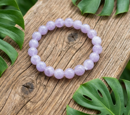 Un bracelet de perles rondes violet clair et translucide est posé sur un morceau de bois texturé. De grandes feuilles de Monstera vertes l'encadrent, créant une ambiance naturelle et apaisante. L'image dégage une impression de pureté et de calme.