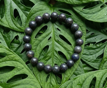 Ce bracelet circulaire est composé de perles rondes et lisses d'un noir profond, parsemées de minuscules inclusions pailletées multicolores évoquant une poussière d'étoiles. Le bijou est posé à plat au centre d'une grande feuille de Monstera verte et brillante, dont les découpes naturelles font ressortir l'éclat mystérieux des pierres.