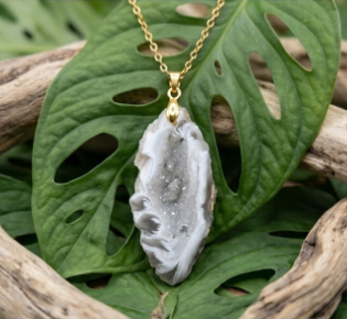 Cette image propose un plan rapproché centré sur la face interne du pendentif. Le pendentif : Il s'agit d'une druse d'agate de forme ovale et organique. Le cœur de la pierre est une cavité tapissée de micro-cristaux scintillants, allant du gris clair au blanc transparent, rappelant une formation de quartz ou une géode ouverte. Le pourtour est formé par des bandes d'agate aux motifs ondulés et fluides dans des tons dégradés de blanc, gris perle et gris ardoise. La pierre est sertie d'un bélière et suspendue à une fine chaîne argentée. L'arrière-plan : Le pendentif repose directement sur de larges feuilles de Monstera d'un vert profond, caractérisées par leurs découpes naturelles typiques (phénotype ajouré). Il est encadré par des morceaux de bois flotté aux tons gris clair, beige et bruns, dont les textures sont rugueuses et noueuses. L'ensemble est posé sur un support en bois clair vieilli. L'éclairage est doux et naturel, mettant en valeur les contrastes entre le brillant des cristaux et l'aspect mat du bois et du feuillage.