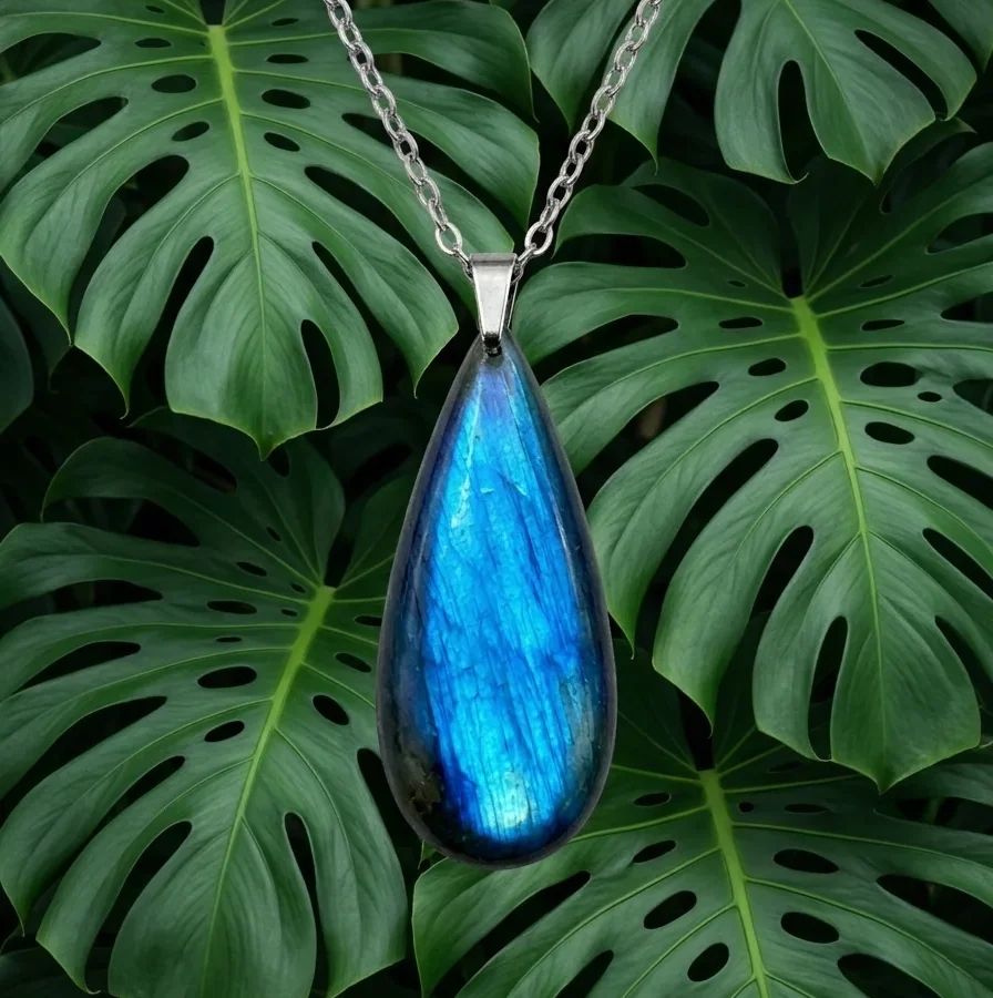 Un pendentif en forme de larme d'un bleu électrique brillant est suspendu au centre d'une composition végétale. Il est entouré de grandes feuilles de monstera et de fougères vert foncé, avec des accents de doré. L'ensemble dégage une ambiance élégante et naturelle sur un fond noir profond.