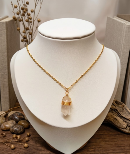 Collier doré arborant un pendentif en cristal de roche transparent, taillé en pointe et entouré d'un fil d'or. Le bijou est présenté sur un buste blanc épuré, sans étiquette de prix. Le décor environnant est composé de bois flotté, de galets lisses et de touches végétales séchées sur un support en bois sombre. L'ensemble dégage une atmosphère naturelle, lumineuse et élégante.