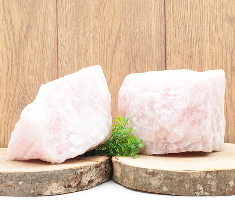 Cette image présente deux morceaux massifs de quartz rose brut, aux formes irrégulières et à la texture cristalline visible. Les pierres, d'une douce teinte rose pâle, reposent sur deux tranches de bois de bouleau naturel, avec de l'écorce sur les côtés. Un petit bouquet de fausse verdure verte est niché entre les deux blocs pour la décoration. Veuillez noter que cette photo est donnée à titre d'exemple et n'est pas contractuelle : la forme, la taille et la couleur exacte des pierres peuvent varier d'une pièce à l'autre.