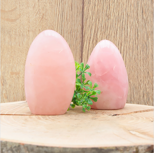 Cette image montre deux morceaux de quartz rose de forme libre, polis et d'une douce couleur rose pâle. La pierre de gauche est sensiblement plus grande que celle de droite, toutes deux reposant sur une tranche de bois de bouleau naturel avec son écorce visible. Un petit bouquet de verdure artificielle est placé discrètement entre les deux blocs pour la décoration. L'ensemble est photographié devant un arrière-plan en bois aux tons chauds, mettant en valeur l'aspect naturel des minéraux.