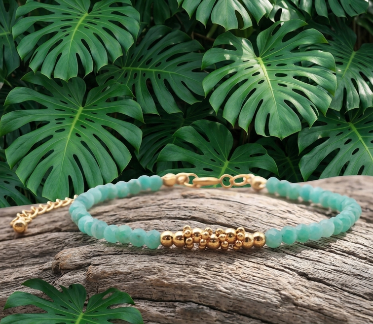 Ce plan rapproché montre un élégant bracelet de perles posé horizontalement sur un morceau de bois flotté gris et texturé. Le bracelet est principalement composé de petites perles turquoise à facettes. Au centre, une grappe de perles sphériques en or brillant ajoute une touche sophistiquée. L'arrière-plan est formé d'un feuillage dense et luxuriant de grandes feuilles de Monstera, créant un décor naturel et tropical.