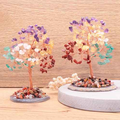Deux petits arbres de vie 7 chakras artisanaux en perles de pierres naturelles colorées sont posés sur un support en bois texturé. Leurs troncs torsadés sont en cuivre doré et reposent sur des bases rocheuses couvertes de petits éclats de pierre. Les perles des arbres sont réparties en grappes de couleurs : bleu, jaune, orange, vert et violet.