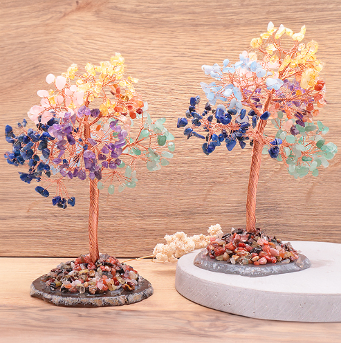 Deux petits arbres de vie 7 chakras artisanaux en perles de pierres naturelles colorées sont posés sur un support en bois texturé. Leurs troncs torsadés sont en cuivre doré et reposent sur des bases rocheuses couvertes de petits éclats de pierre. Les perles des arbres sont réparties en grappes de couleurs : bleu, jaune, orange, vert et violet.
