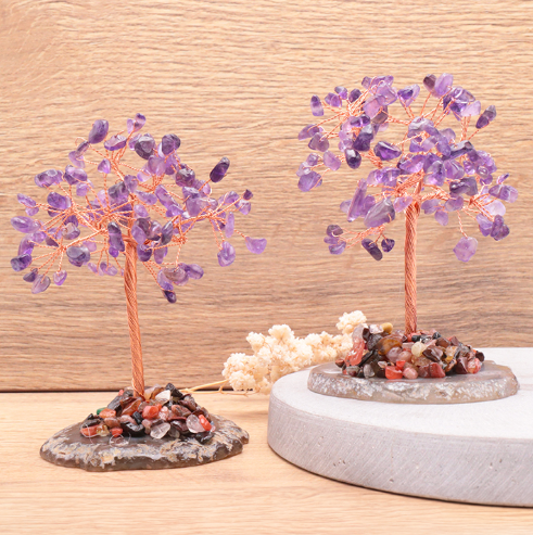 Cette image présente deux arbres de vie améthyste artisanaux, d'environ 10 cm de hauteur, avec des troncs et des branches en fil de cuivre torsadé. Leur feuillage est constitué d'une multitude de petits fragments d'améthyste violette, offrant des reflets éclatants et variés. Chaque arbre repose sur une base naturelle en agate d'une couleur plus sombre. À l'arrière-plan, un mur en bois clair crée une atmosphère naturelle et chaleureuse.