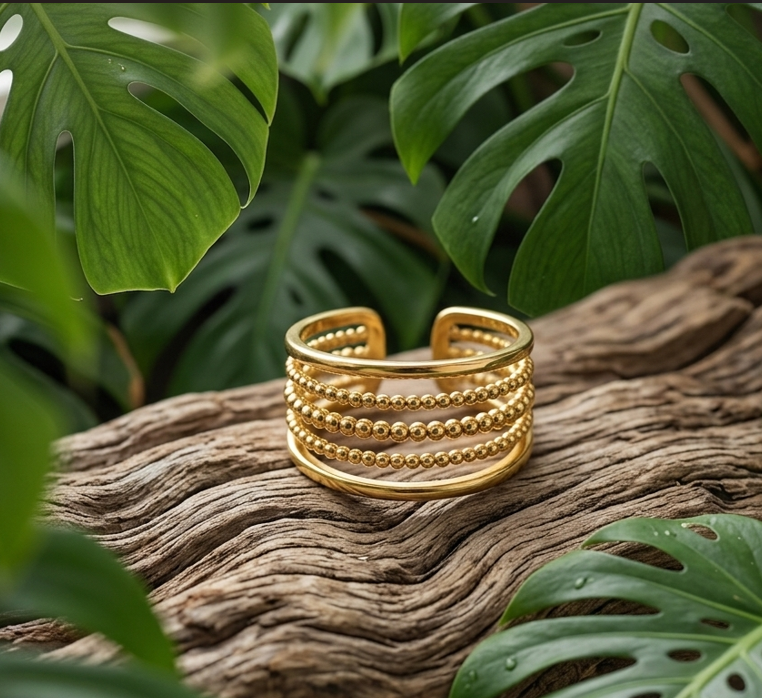 une large bague réglable, de couleur dorée brillante, repose sur une surface de bois flotté très texturée. Elle est composée de cinq anneaux empilés : deux anneaux lisses aux extrémités et trois anneaux texturés de petites perles au centre. Le décor est entouré de grandes feuilles de Monstera vertes et vibrantes, créant une ambiance naturelle et artisanale.
