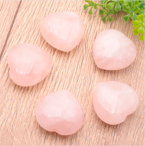 L'image montre cinq pierres de quartz rose taillées en forme de cœurs lisses et bombés, disposées en cercle sur une surface en bois clair. Chaque cœur présente une teinte rose pâle délicate avec de légères variations de transparence et des marbrures naturelles blanchâtres. Une petite branche de verdure vive apparaît dans le coin supérieur droit, apportant un contraste de couleur avec le rose tendre des minéraux. L'ensemble dégage une atmosphère douce et apaisante, mettant en valeur la texture polie et brillante des cristaux.