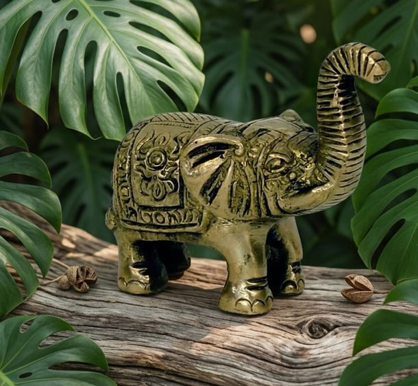 Une statuette d'éléphant en laiton doré est posée sur une branche de bois flotté. L'animal, finement ciselé avec des motifs décoratifs sur le dos, se tient debout, la trompe fièrement relevée. L'ensemble est mis en valeur par un arrière-plan composé de larges feuilles de Monstera d'un vert profond.