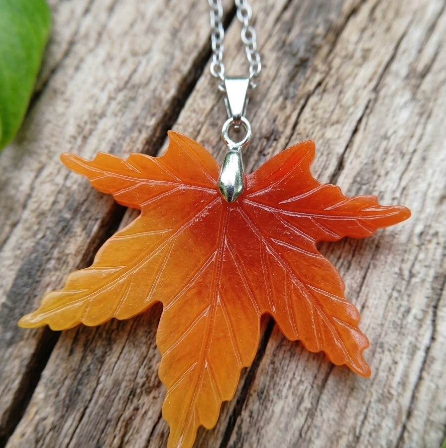 Un pendentif en forme de feuille d'érable, sculpté dans une coquille aux nuances flamboyantes allant de l'orange vif au jaune ambré, est présenté en gros plan. Il est suspendu à une fine chaîne argentée et repose sur une pièce de bois brut aux rainures marquées. Quelques feuilles de Monstera vert éclatant apparaissent délicatement sur les bords de l'image.