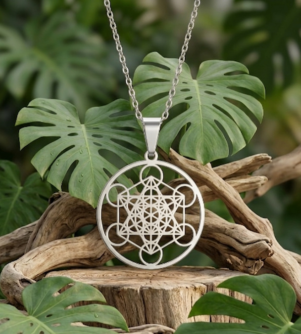 e collier argenté porte un large pendentif circulaire et ajouré représentant le Cube de Mététron, un motif géométrique sacré complexe composé de cercles et de lignes entrelacés. Il est présenté dans un décor naturel sur un socle en bois brut, entouré de branches sèches et de grandes feuilles de Monstera vertes.