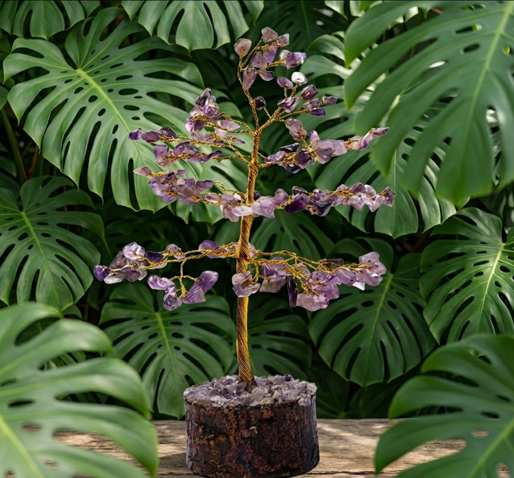 Un arbre de vie miniature, composé de fils de cuivre dorés et torsadés, est orné exclusivement de petites pierres d'améthyste violette aux éclats translucides. L'arbre est solidement ancré sur sa base d'origine : un socle cylindrique en bois sombre et brut, à l'écorce rugueuse, recouvert sur le dessus de petits éclats d'améthyste. L'ensemble est posé sur une table en bois clair. À l'arrière-plan et au premier plan sur les côtés, un décor dense de grandes feuilles vertes et découpées de Monstera encadre délicatement l'objet, créant une ambiance végétale et harmonieuse.