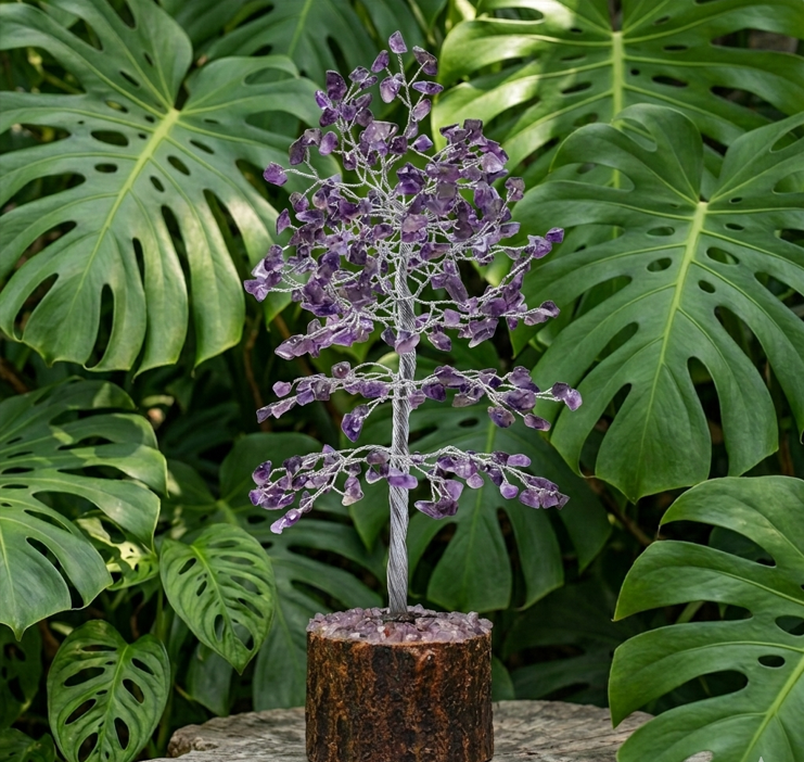 Un arbre de vie composé de fils de cuivre dorés et torsadés, est orné exclusivement de petites pierres d'améthyste violette aux éclats translucides. L'arbre est solidement ancré sur sa base d'origine : un socle cylindrique en bois sombre et brut, à l'écorce rugueuse, recouvert sur le dessus de petits éclats d'améthyste. L'ensemble est posé sur une table en bois clair. À l'arrière-plan et au premier plan sur les côtés, un décor dense de grandes feuilles vertes et découpées de Monstera encadre délicatement l'objet, créant une ambiance végétale et harmonieuse.