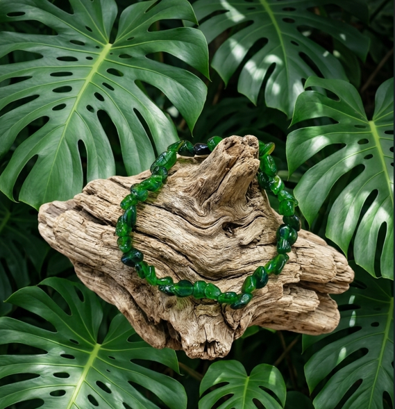 Cette image met en valeur un bracelet composé de petites pierres roulées d'un vert émeraude intense, délicatement posé sur un morceau de bois flotté très texturé et noueux. L'ensemble est harmonieusement entouré par un arrière-plan luxuriant de grandes feuilles de monstera vert vif.