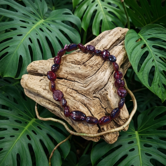 Cette image met en scène un bracelet composé de perles de pierres roulées d'un rouge bordeaux profond et brillant, disposé sur un bloc de bois flotté beige très texturé. En arrière-plan, le décor est composé d'un feuillage dense et sombre de grandes feuilles de monstera.