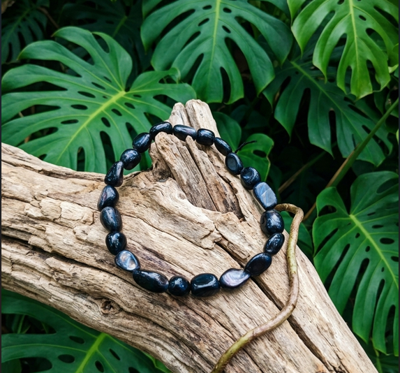 Cette image en gros plan montre un bracelet composé de perles de pierres noires irrégulières et polies. Le bracelet est posé sur un morceau de bois flotté texturé, dont les fibres sèches contrastent avec la surface brillante des pierres. Derrière le bois flotté se trouve une plante de Monstera, dont les grandes feuilles vertes et découpées remplissent l'arrière-plan.