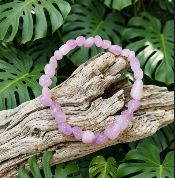 Cette photographie met en scène un bracelet élastique composé de perles de pierres naturelles polies d'un rose violacé doux. Le bracelet est posé en premier plan sur un morceau de bois flotté texturé et blanchi, l'ensemble étant entouré par un arrière-plan dense de grandes feuilles vertes et découpées de Monstera.