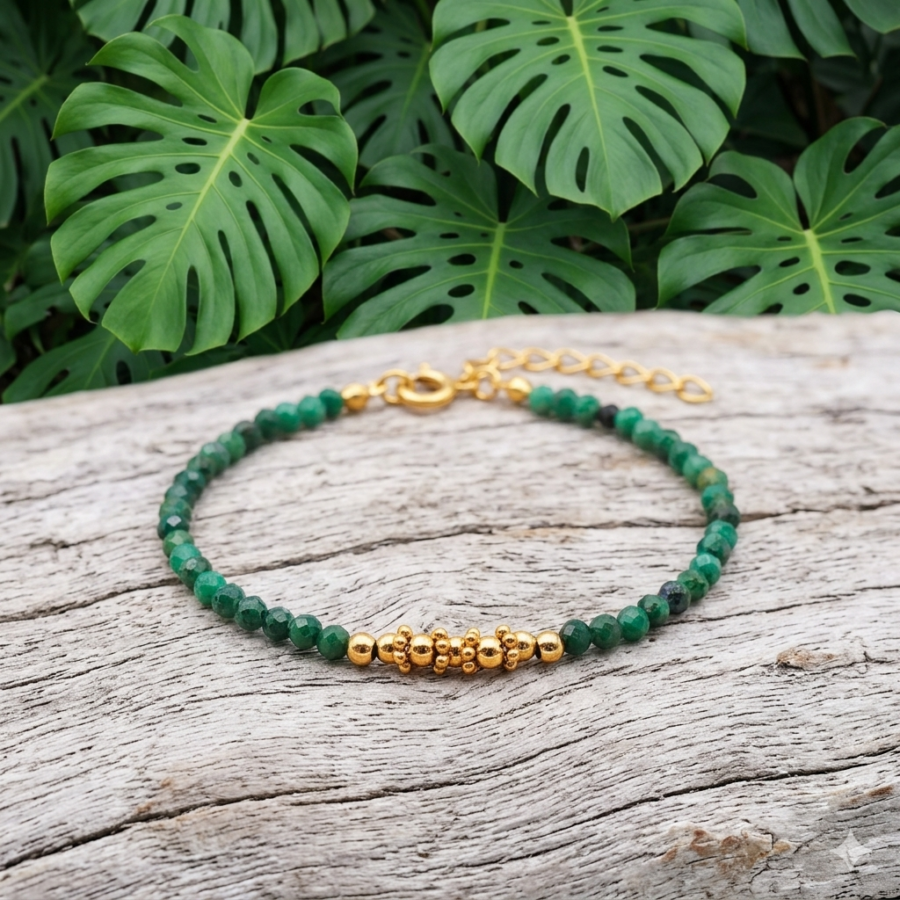 Bracelet en perles de Quartz Fuchsite aux facettes vert émeraude et éléments dorés, disposé sur du bois flotté. À l'arrière-plan, de grandes feuilles de Monstera apportent une ambiance tropicale et apaisante. Le contraste entre le bois grisâtre et le vert éclatant des pierres souligne la finesse du bijou.